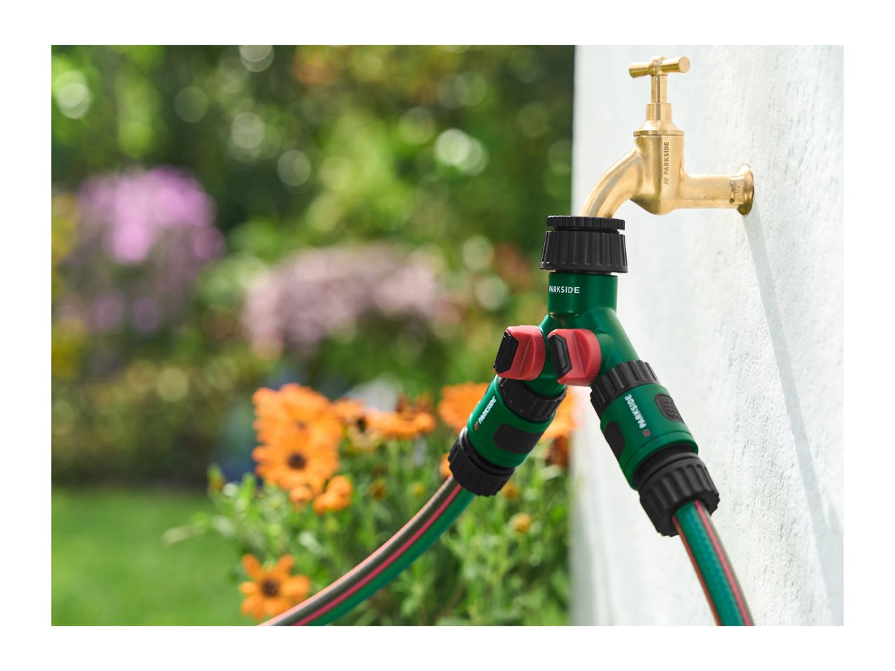 Garden tap with a two-way hose splitter and two green garden hoses, surrounded by flowers.