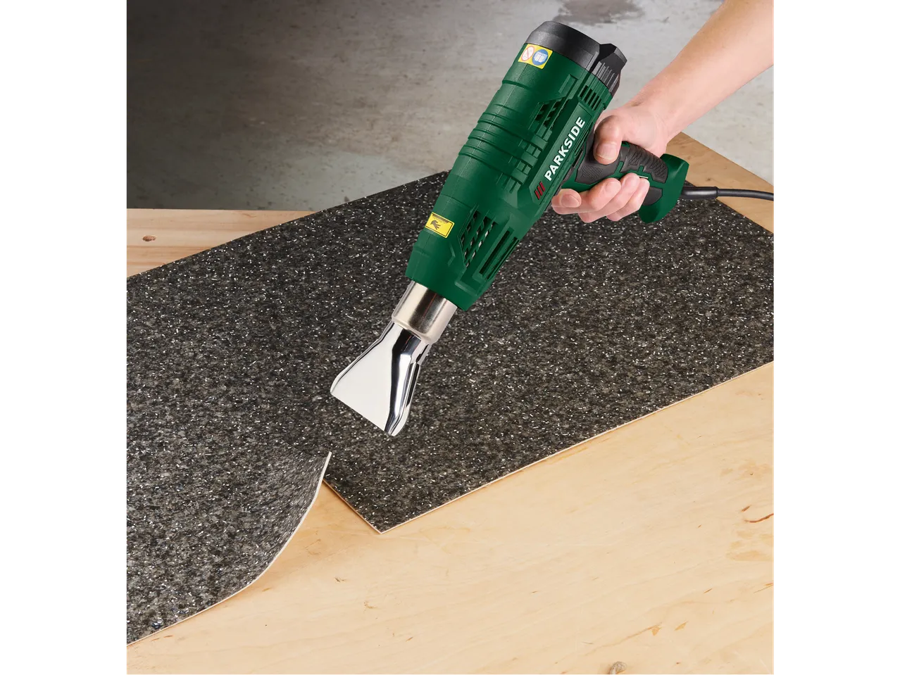 Roka drži zeleno toplotno pištolo, ki segreva temno, teksturirano površino na leseni deski.