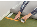 Mãos aplicando fita adesiva 3M em base verde junto à parede.