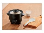 Black rice cooker with cooked rice, a measuring cup of uncooked rice, and a serving spoon.
