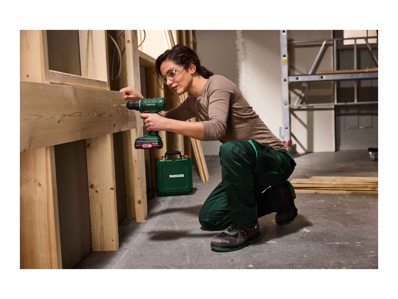 Woman in safety glasses and workwear using a cordless drill on a wooden frame.