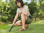 Eine Frau hockt im Garten und benutzt ein grünes Handgerät auf dem Rasen.