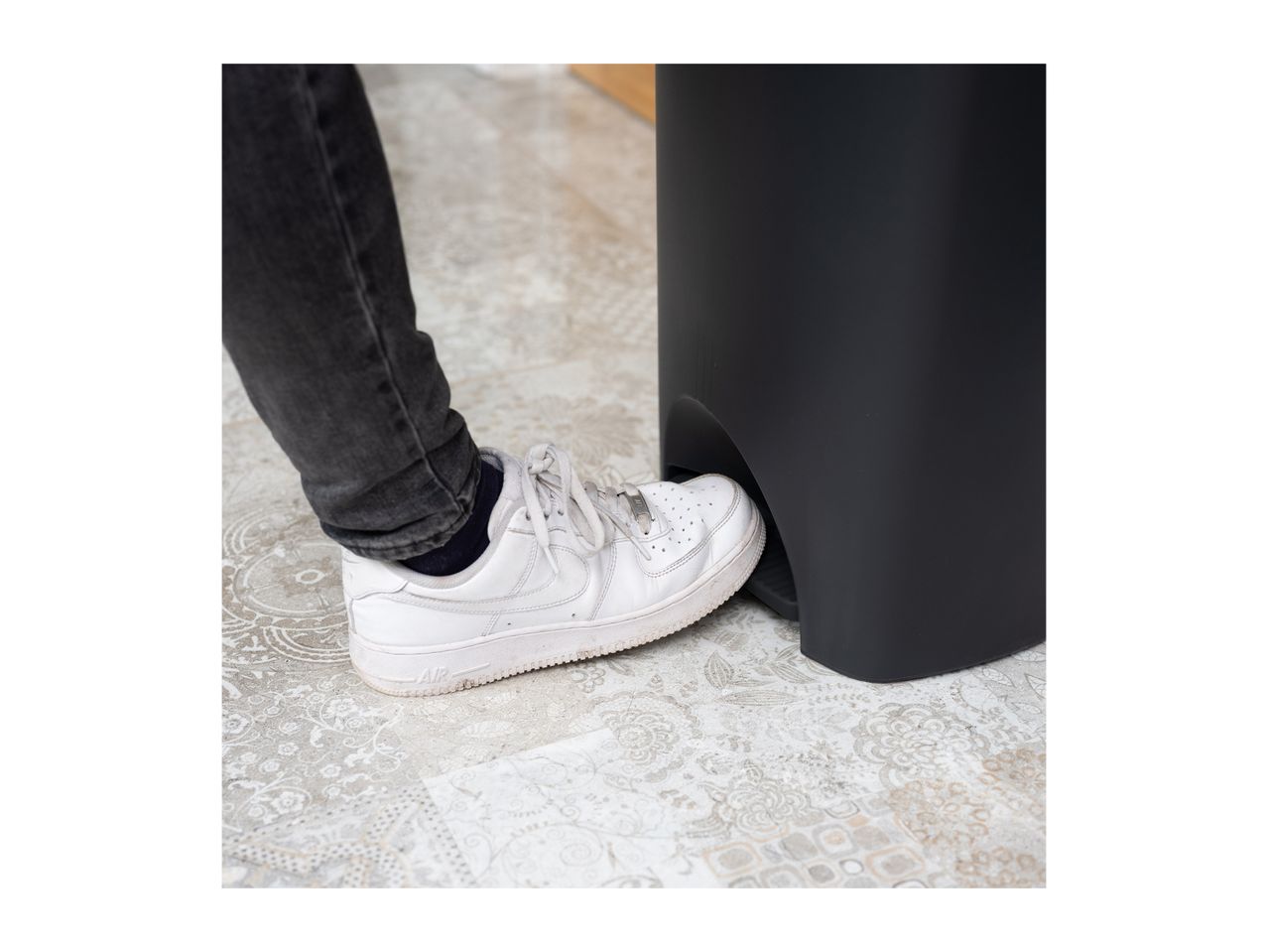 A person's foot in a white sneaker pressing the pedal of a black pedal bin.