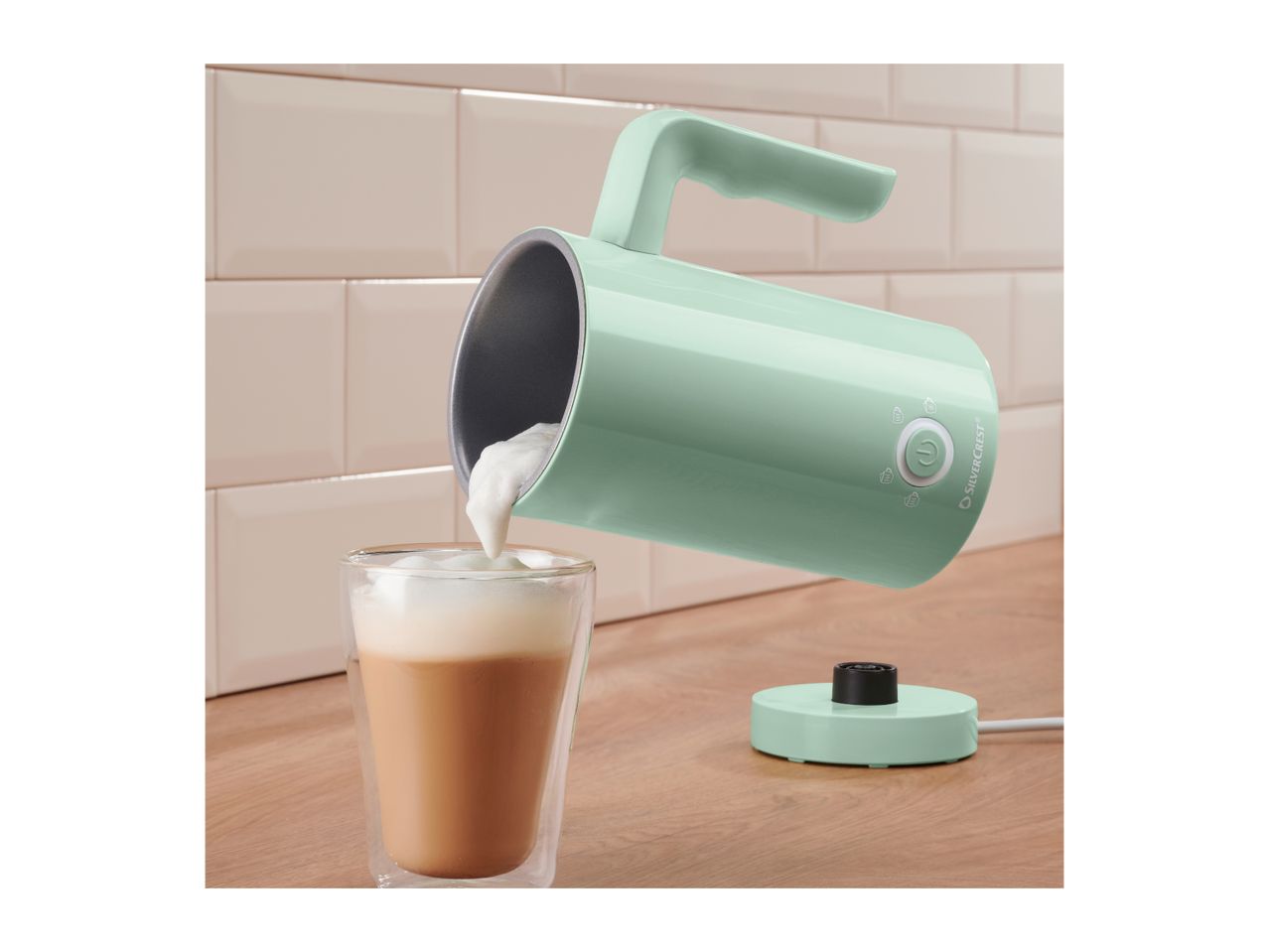 Mint green milk frother pouring foam into a glass of coffee on a wooden counter.