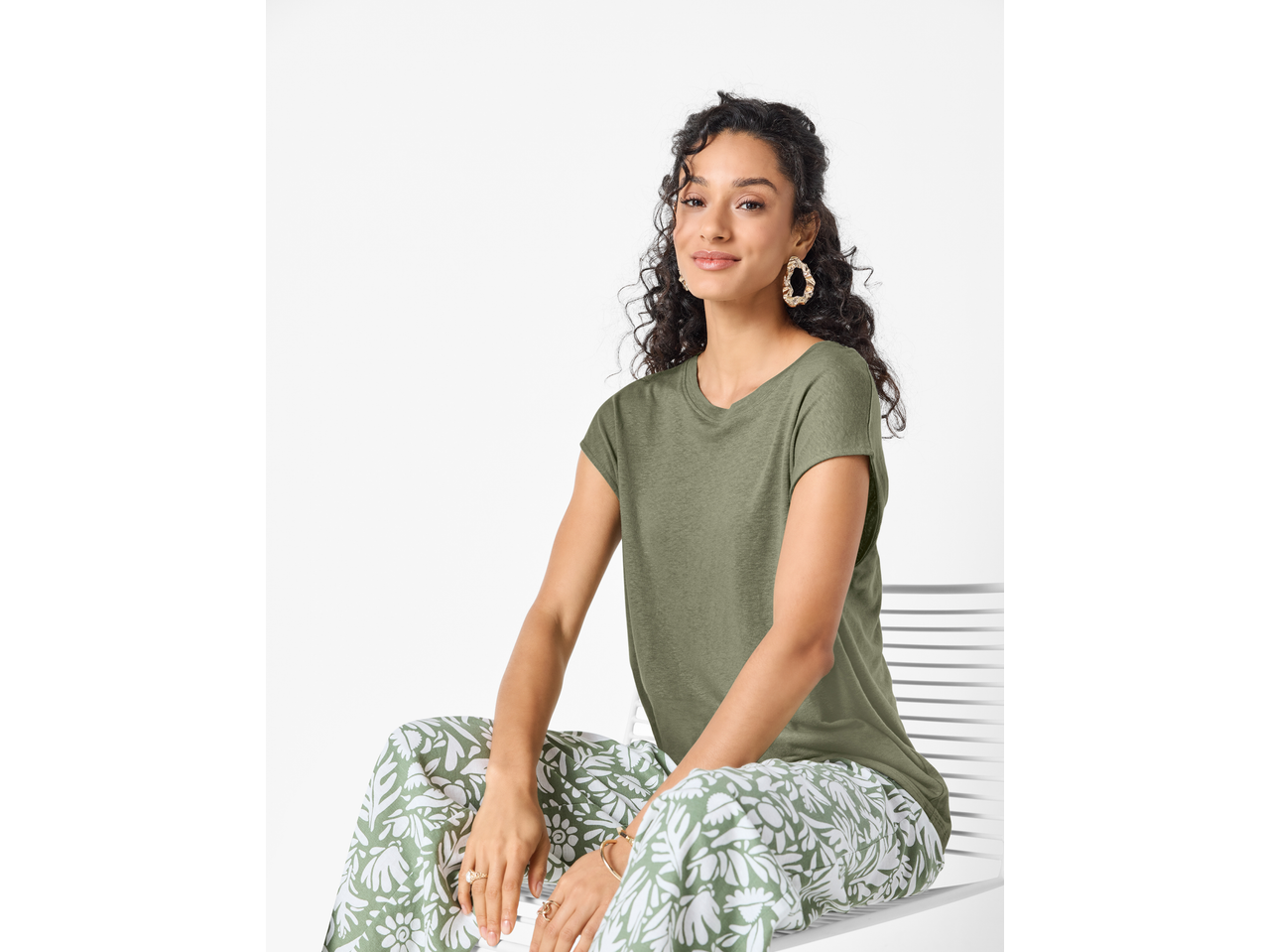 Mulher com t-shirt verde-oliva e calças com estampado floral branco, sentada e sorrindo.