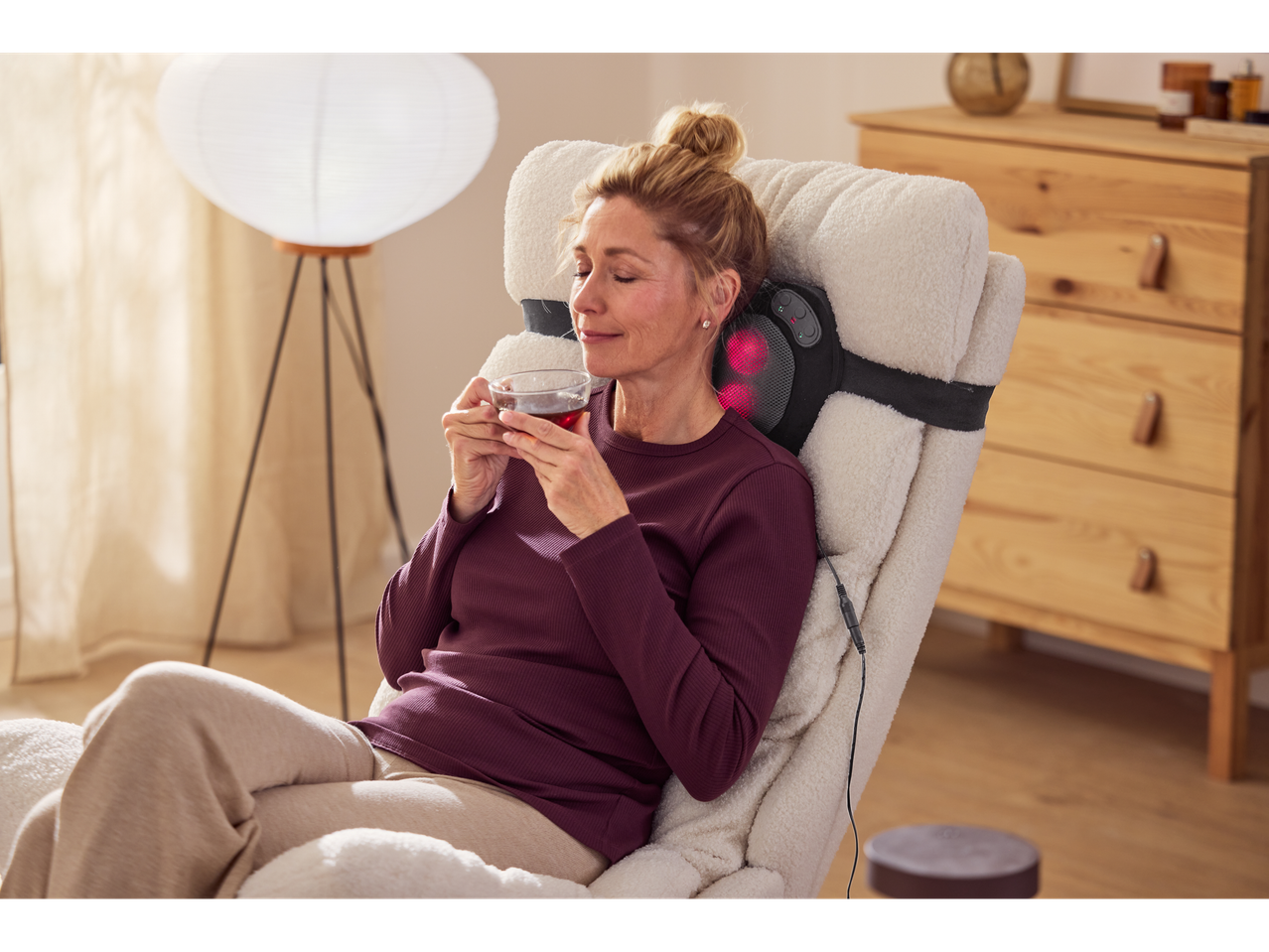 Žena se opušta u fotelji sa SENSIPLAST® Shiatsu masažnim jastukom i šoljom čaja.