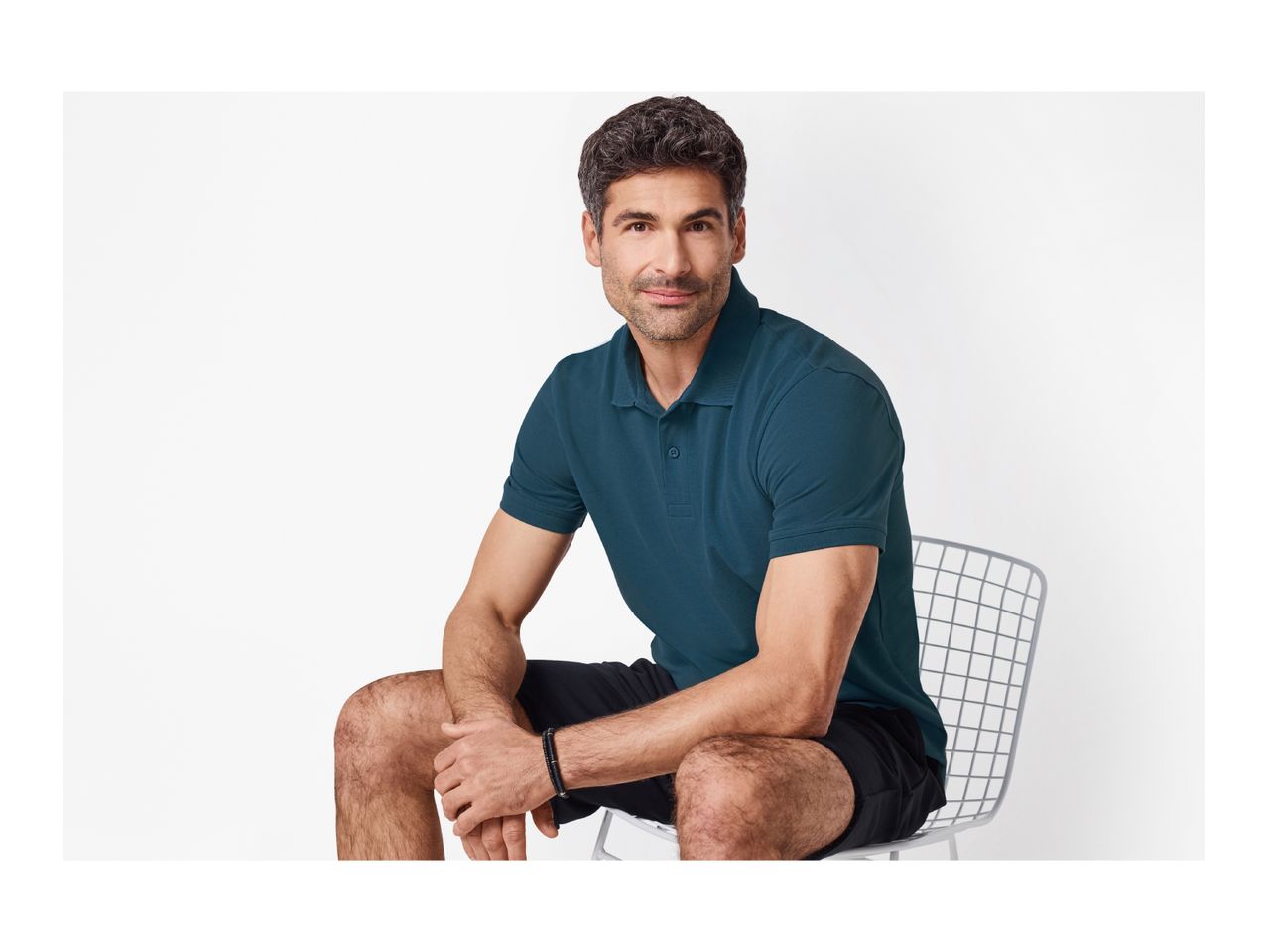 Man in a dark teal polo shirt and black shorts, sitting on a white chair.