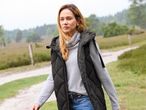 Egy nő szürke garbóban, fekete steppelt mellényben és farmerben áll egy ösvényen.