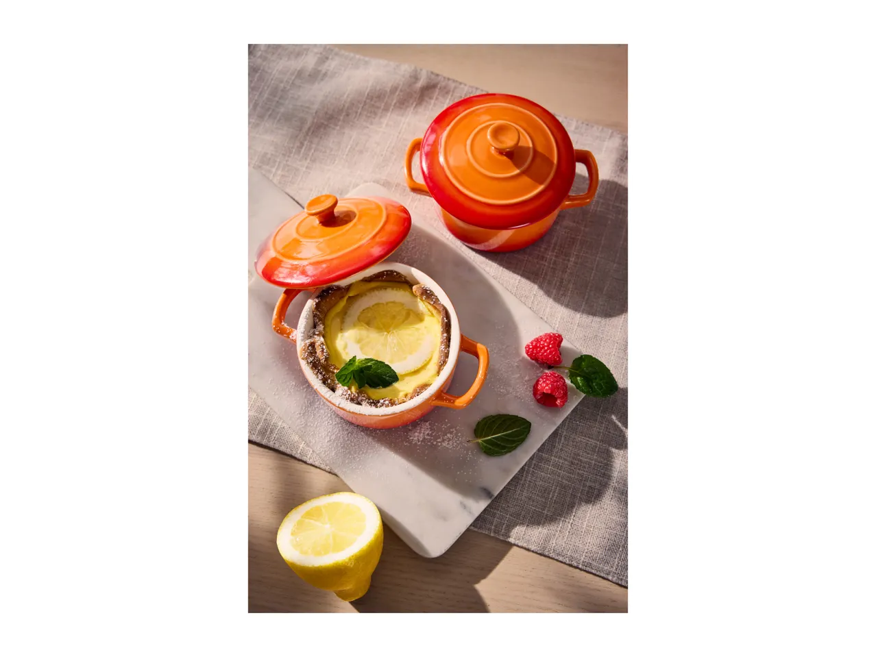 Lemon dessert in an orange ceramic pot, with raspberries and mint on a marble board.