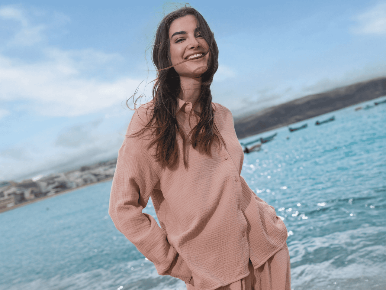 Lachende Frau in rosa Hemd und Hose am Strand mit Meer und Bergen im Hintergrund.