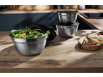 Stainless steel mixing bowls with lids, one filled with salad, on a wooden kitchen counter.