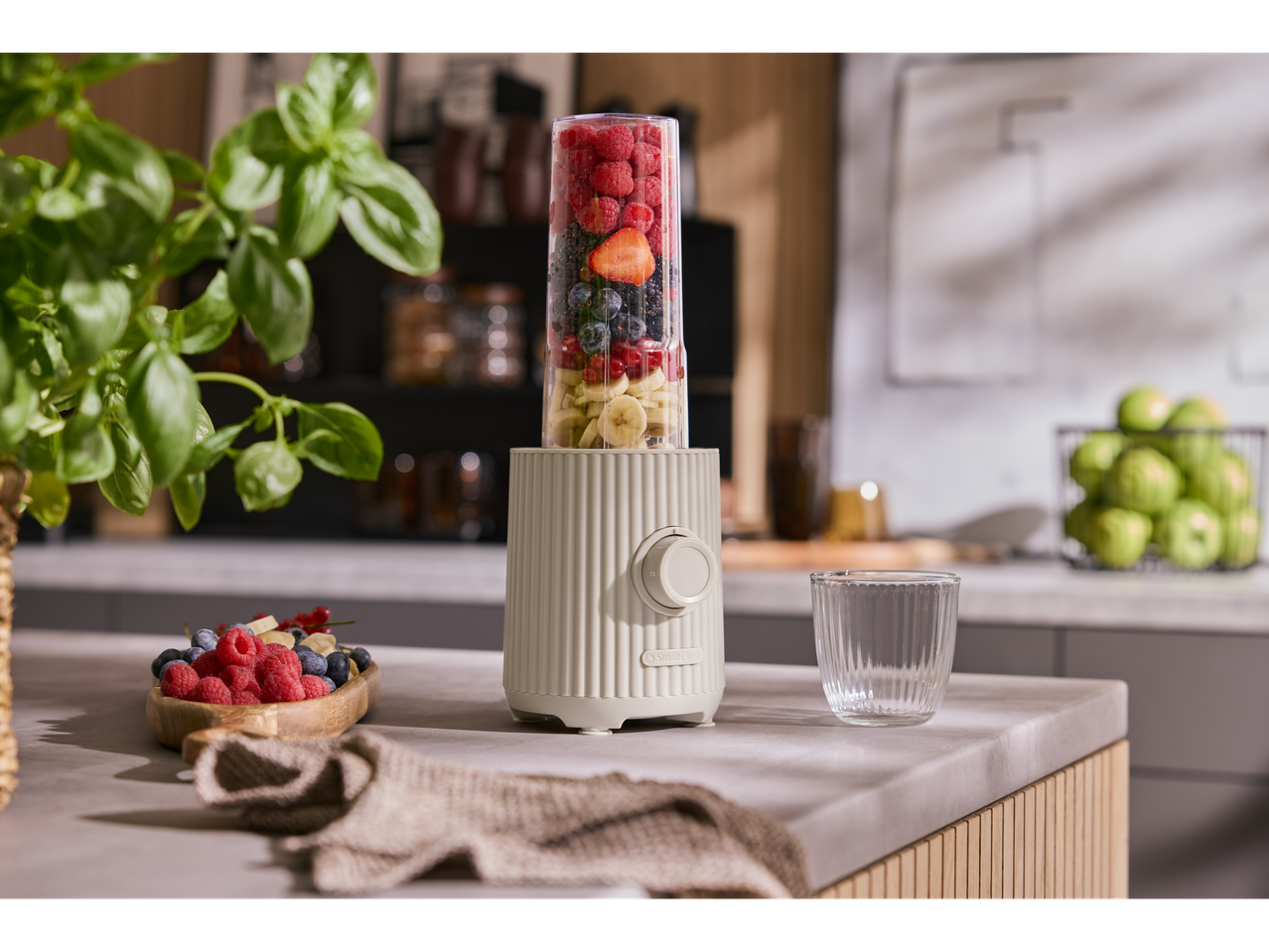 Blender cu fructe de pădure și banană pe blatul de bucătărie cu busuioc și fructe.