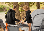 Casal preparando café em uma mesa dobrável de camping perto de uma barraca.