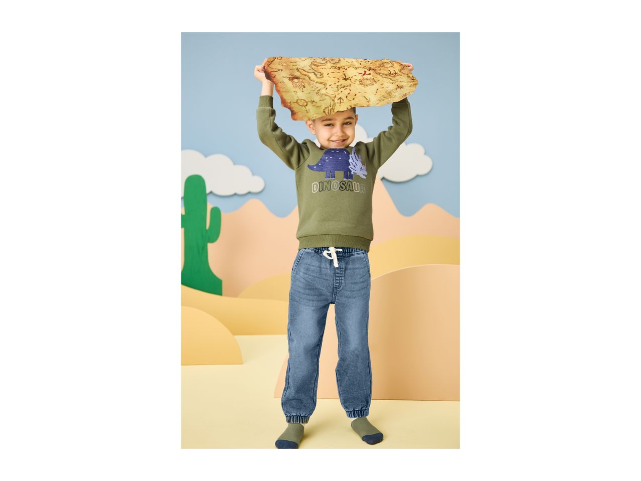 Boy in a green dinosaur sweatshirt and blue jeans holding a treasure map.