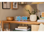 A modern LED digital clock sits on a wooden table, surrounded by books and plants.