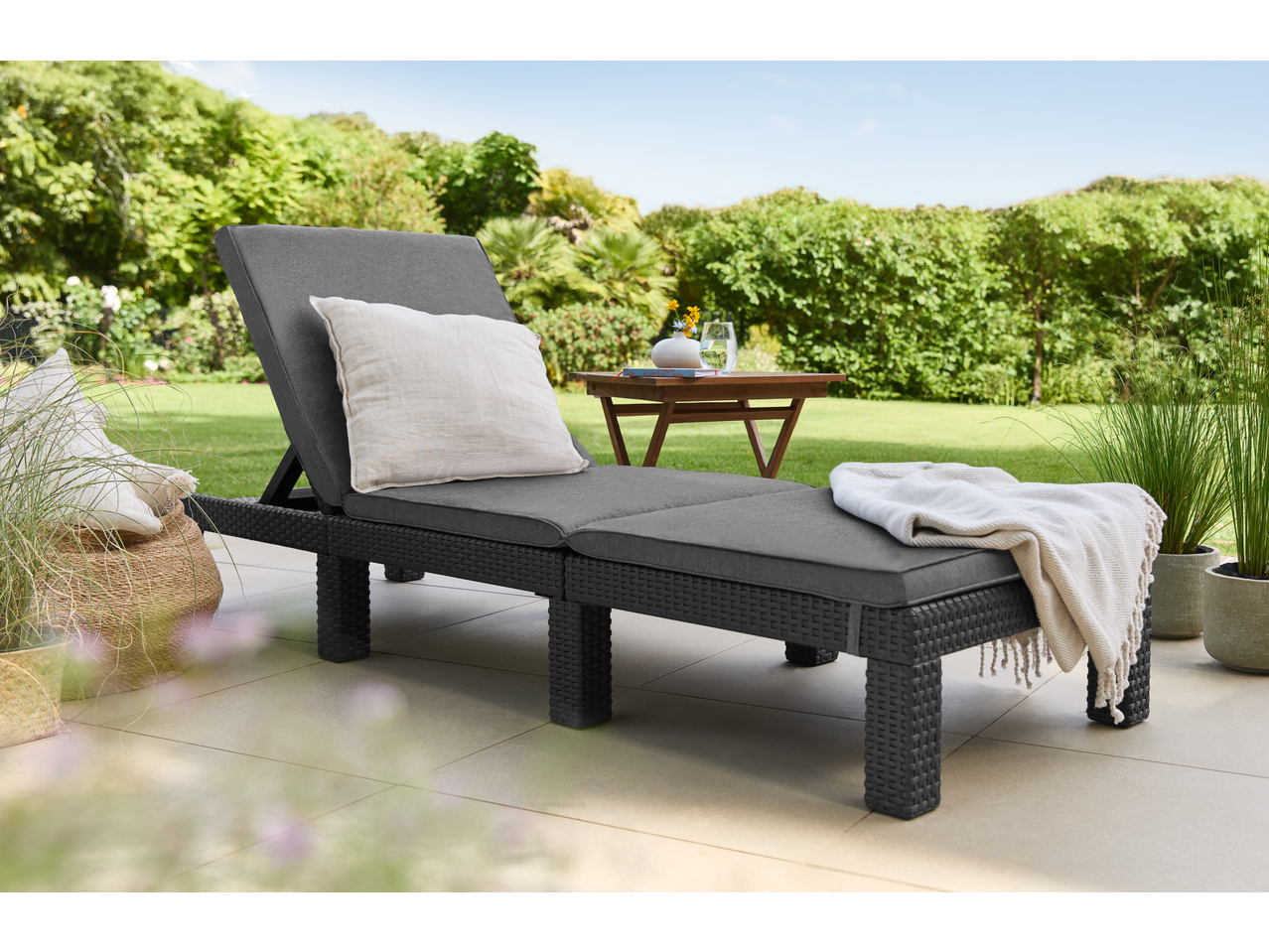 Livarno Wicker Sun Lounger with grey cushions and white pillow on a patio with garden background.