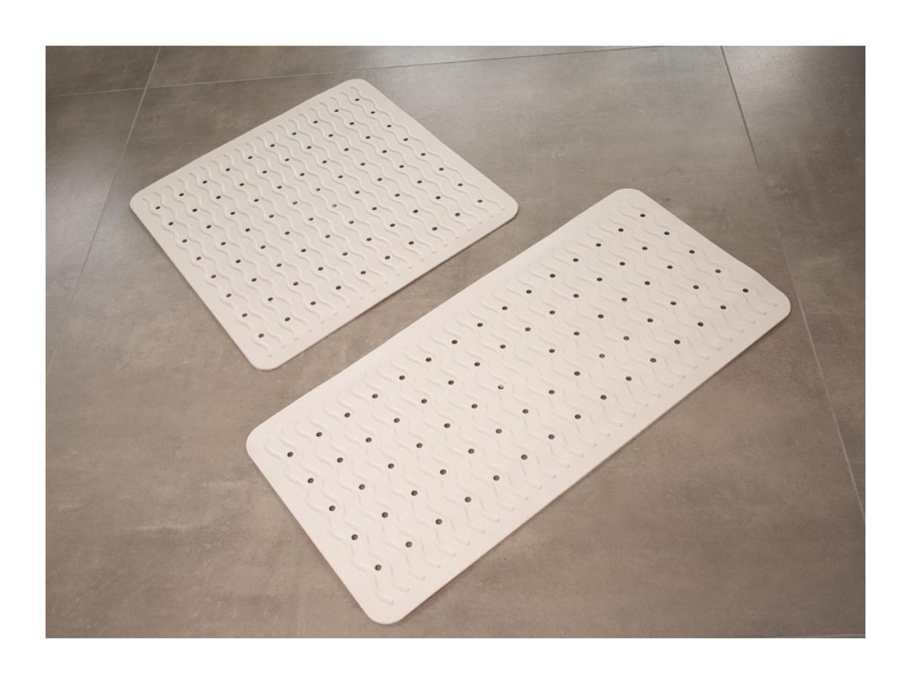 Two white bath mats with wavy patterns and drainage holes on a gray tiled floor.