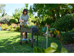 Femeie udând o jardinieră înălțată cu un temporizator de apă într-o grădină însorită.