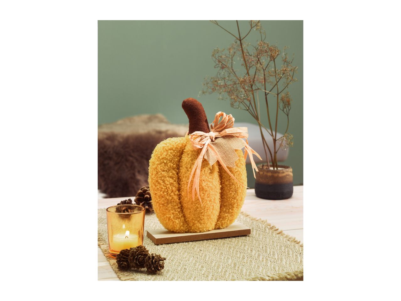 A fluffy orange pumpkin decoration with a brown stem and raffia bow, surrounded by fall decor.