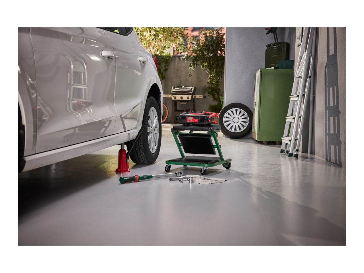 A car jacked up in a garage with a torque wrench, rolling stool, and charger nearby.