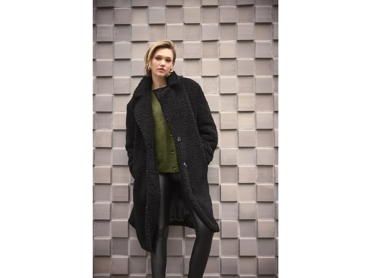 Žena u Esmara kardiganu i crnom teddy kaputu ispred teksturiranog zida.