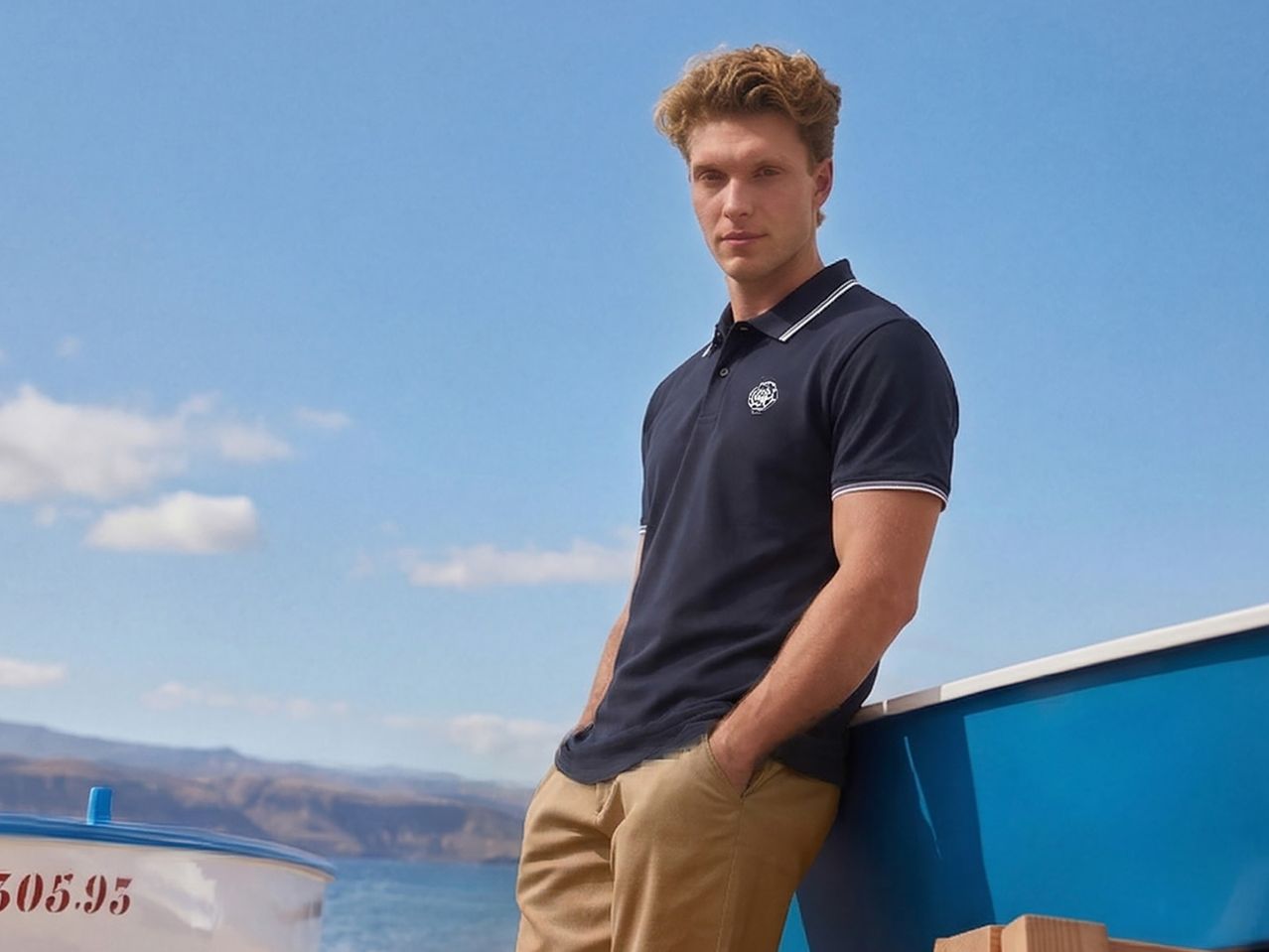 Man in a navy polo shirt and khaki trousers, leaning on a boat by the sea.