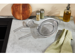 Three stainless steel strainers on a kitchen counter with a wooden board, spices, and olive oil.