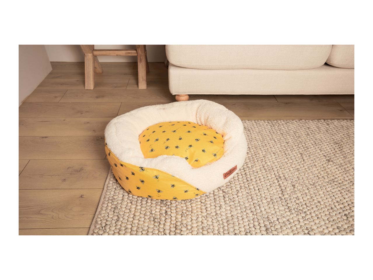 Yellow pet bed with bee print and cream fleece lining on a rug next to a sofa.