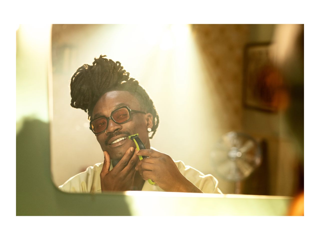 Man with dreadlocks and glasses shaving with an electric shaver in a mirror.