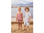 Two children in muslin summer outfits on a beach with the sea and city in the background.