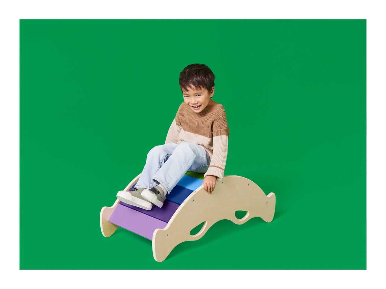 A smiling child sits on a colorful wooden rocker against a green background.