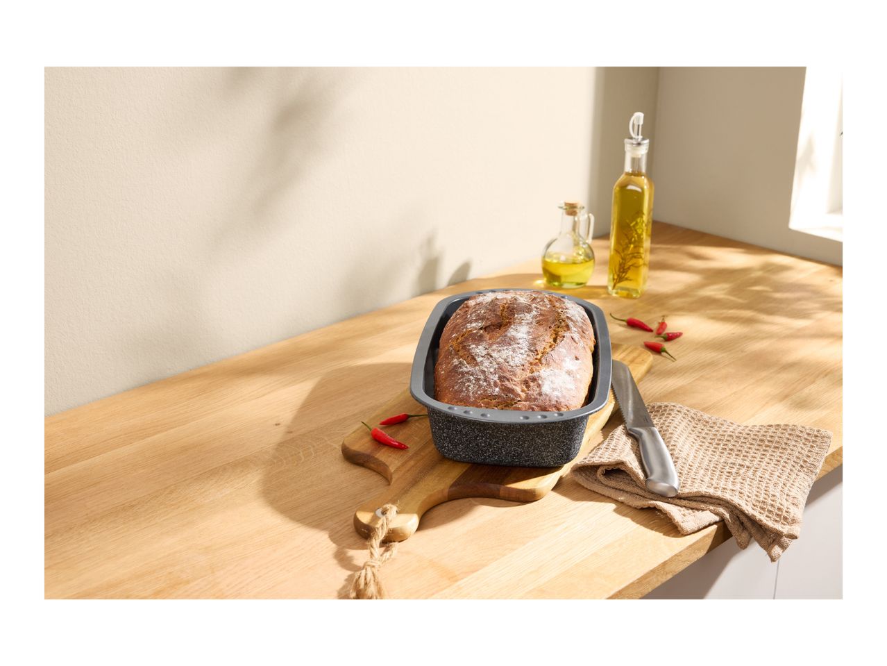 Freshly baked bread in a loaf pan on a wooden cutting board with olive oil and chilies.
