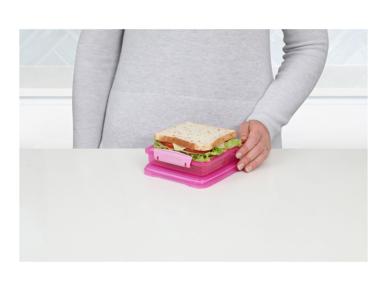 A person holding a sandwich in a pink lunchbox on a white table.