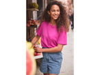 Uma mulher sorridente com uma camiseta rosa e shorts jeans segura uma caixa de maçãs.