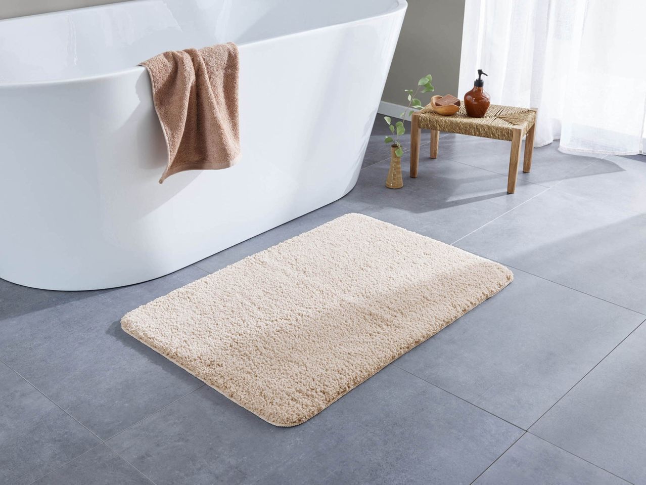 Un tappeto da bagno beige su un pavimento piastrellato grigio accanto a una vasca da bagno bianca.