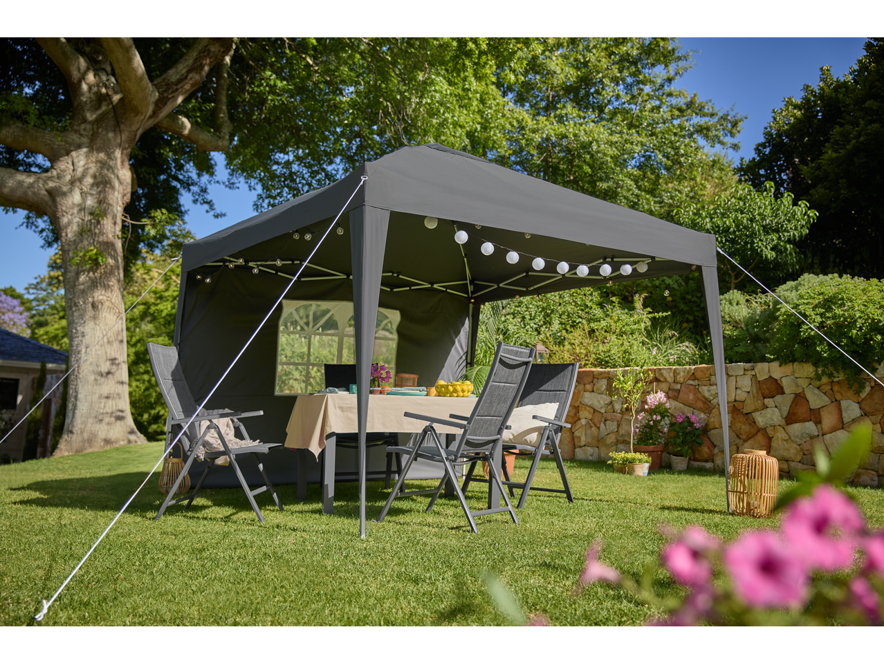 Grey gazebo with string lights and outdoor furniture on a sunny lawn.