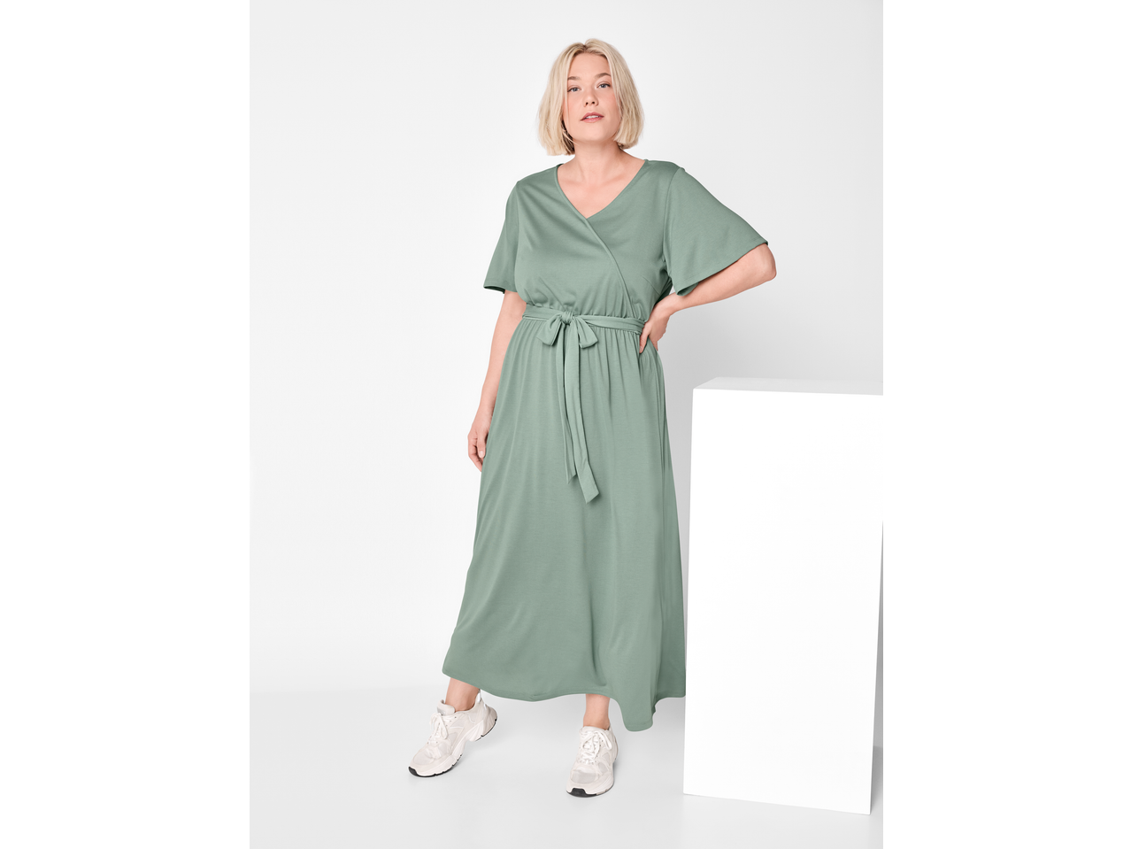 Žena u dugoj zelenoj ESMARA haljini s remenom i tenisicama.