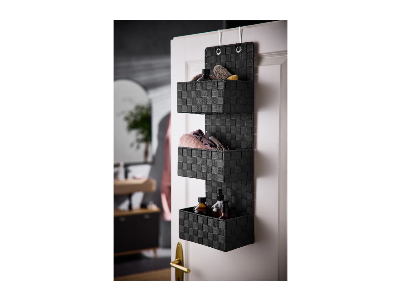 Black woven over-the-door organizer with three baskets holding toiletries and towels.