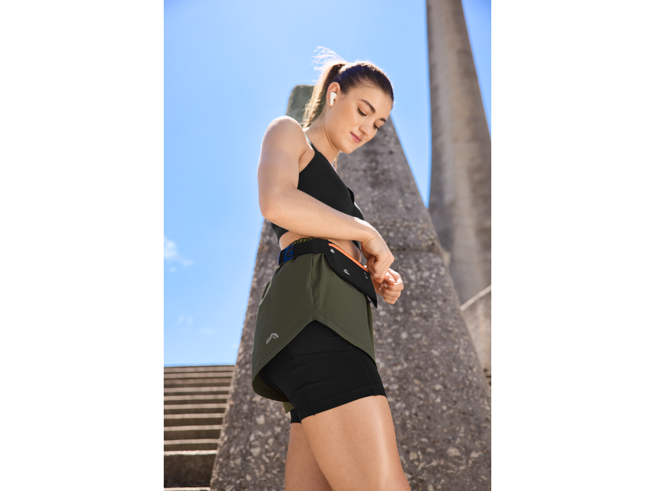 Woman in a black sports bra and olive green running shorts with a fanny pack.