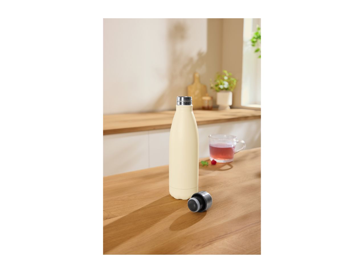Insulated stainless steel bottle on a wooden table next to its lid and a cup of tea.