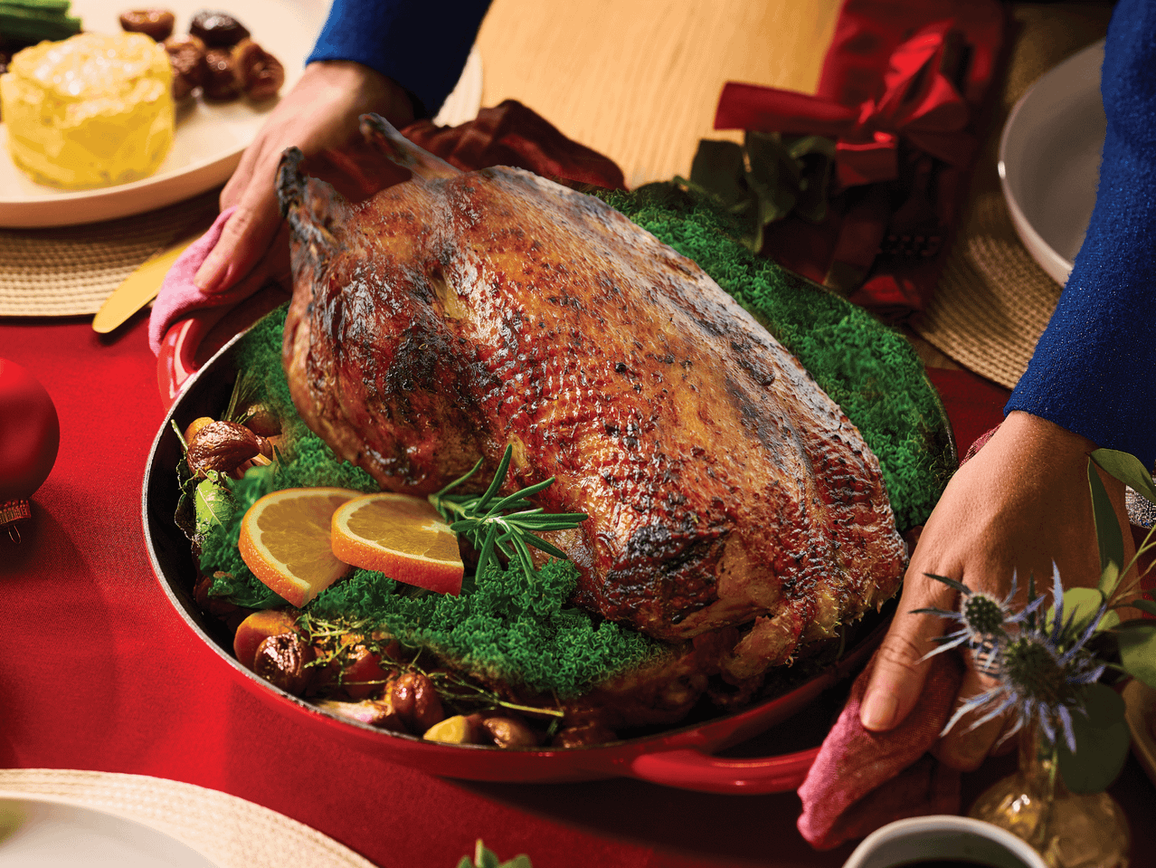 Eine Person serviert eine knusprig gebratene Ente, garniert mit Grünkohl, Orangen und Rosmarin, auf einem festlich gedeckten Tisch.