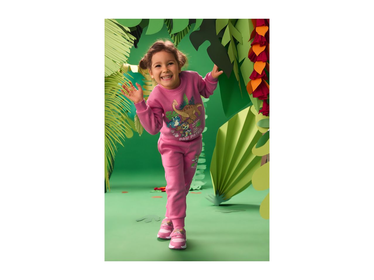 Smiling girl in a pink dinosaur-themed tracksuit and sneakers, surrounded by green paper leaves.