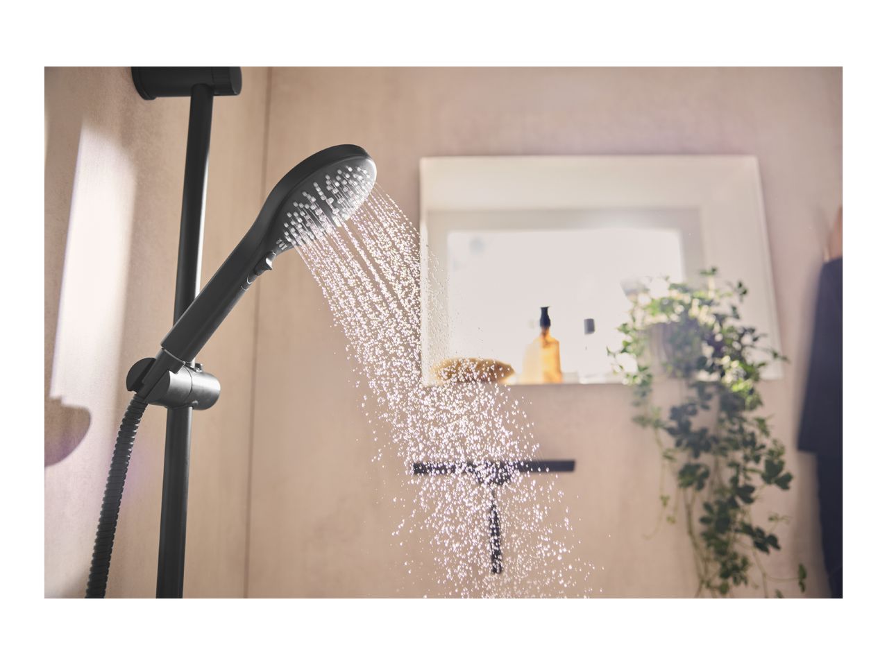 Black shower head spraying water in a modern bathroom with a mirror and plant.