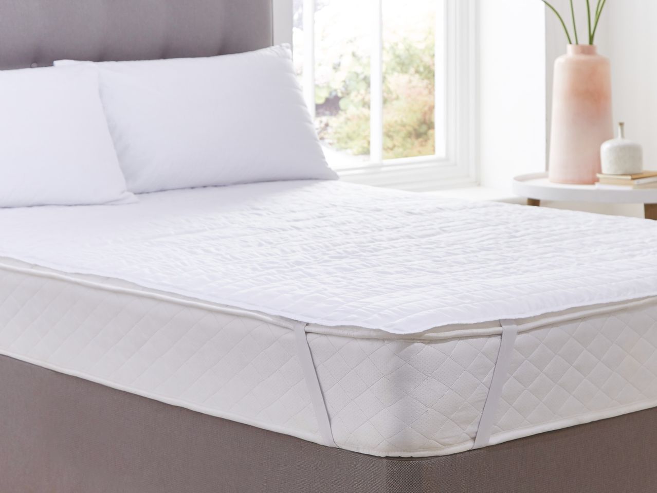 White quilted mattress protector on a bed with pillows, next to a window