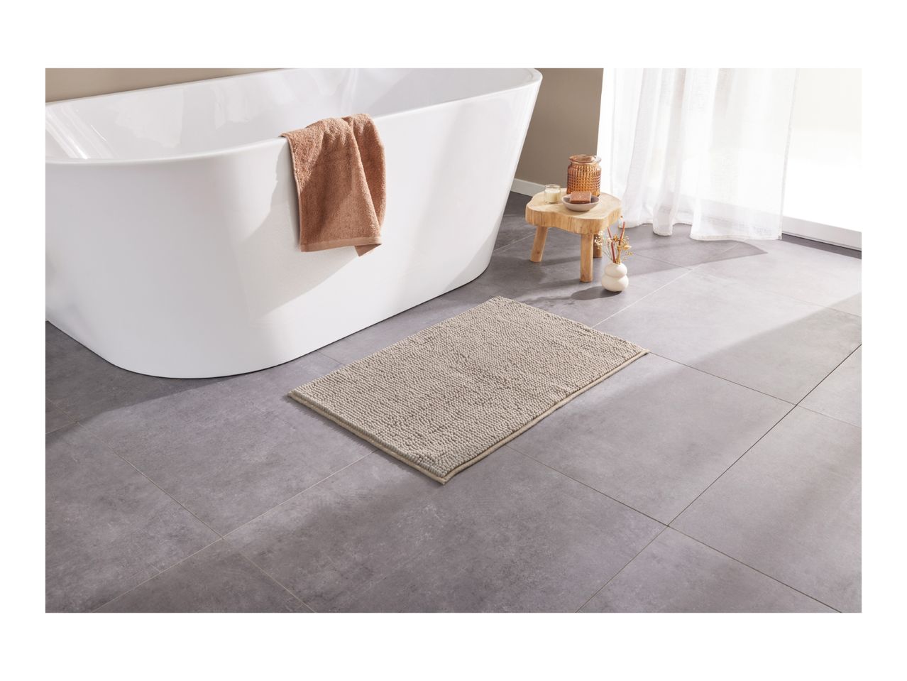 A modern bathroom with a white freestanding bathtub, a beige bath mat, and a wooden stool.