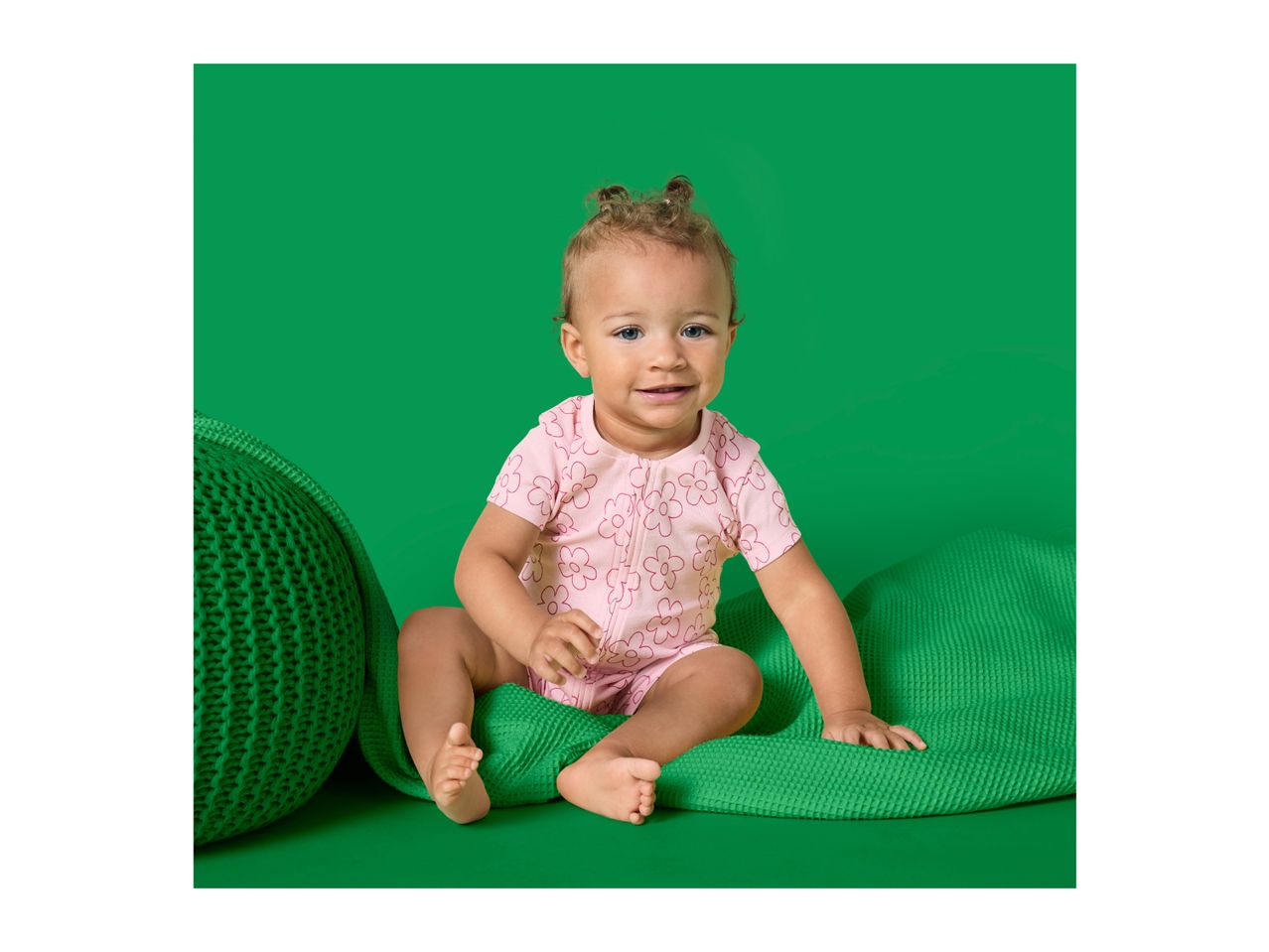 A baby in a pink floral romper sitting on a green blanket with a green pouf.