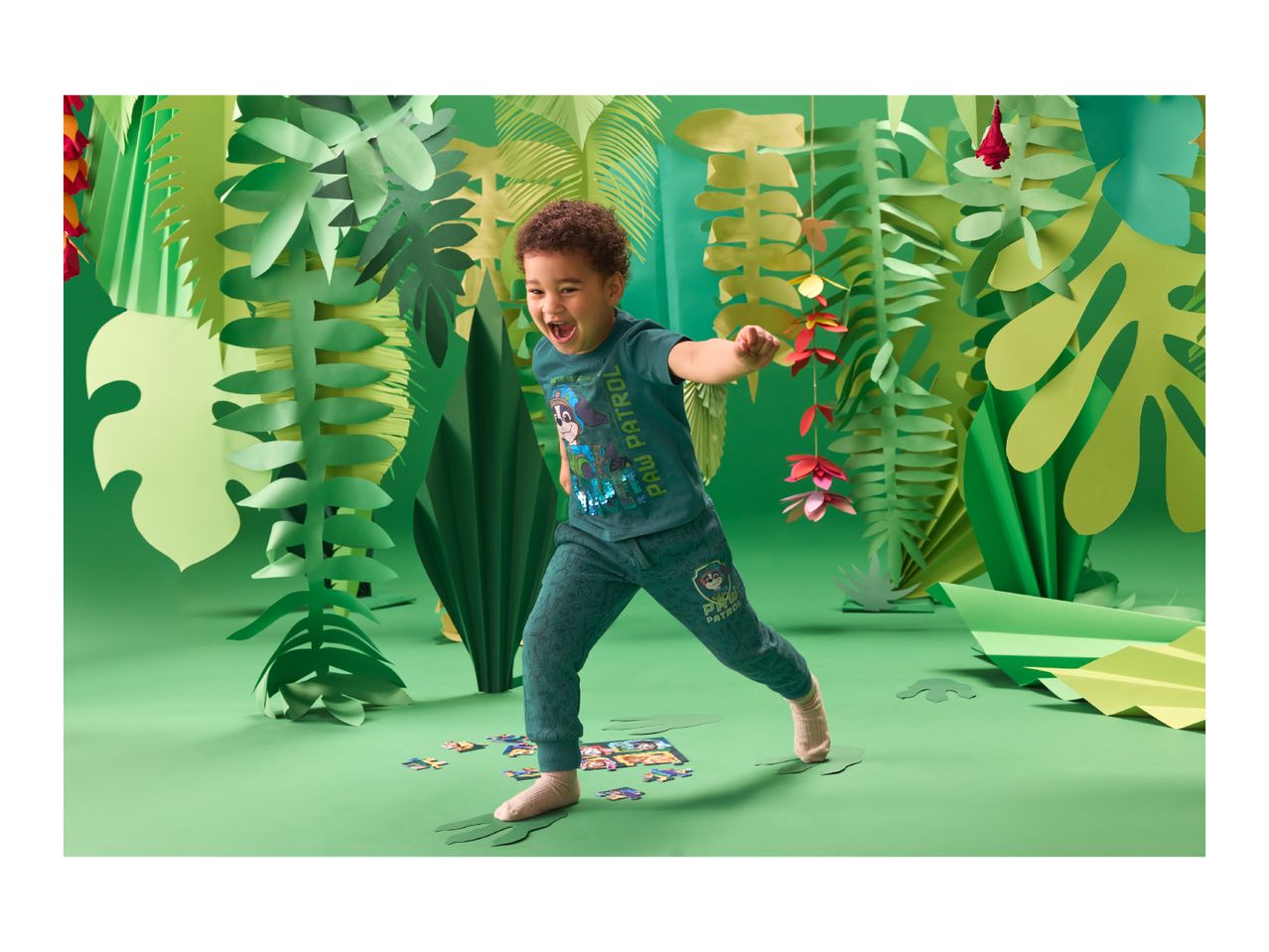A boy in a green Paw Patrol t-shirt and joggers, playing with a puzzle in a jungle setting.