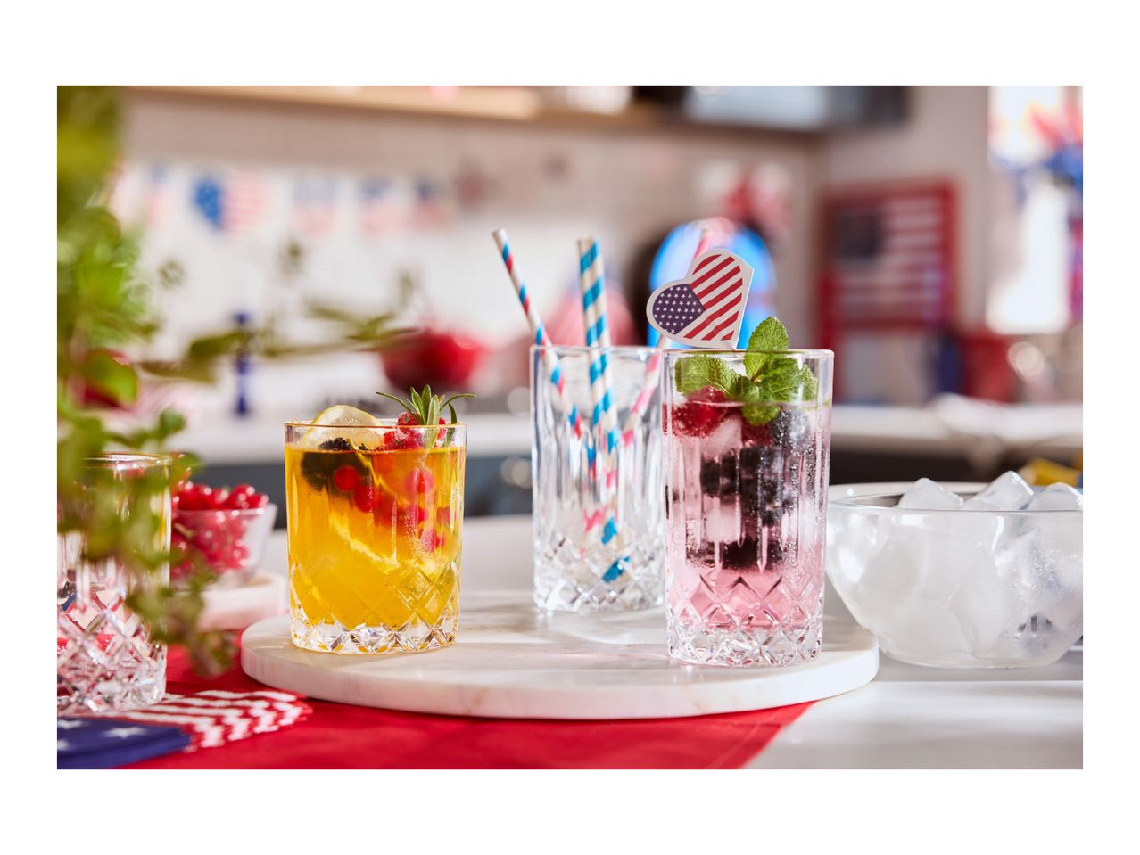Two festive drinks with fruit and ice, one with a US flag pick, on a red tablecloth.