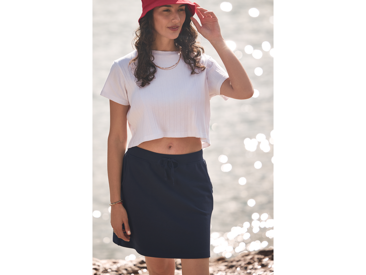 Model purtând un tricou alb și o fustă sport bleumarin lângă mare.