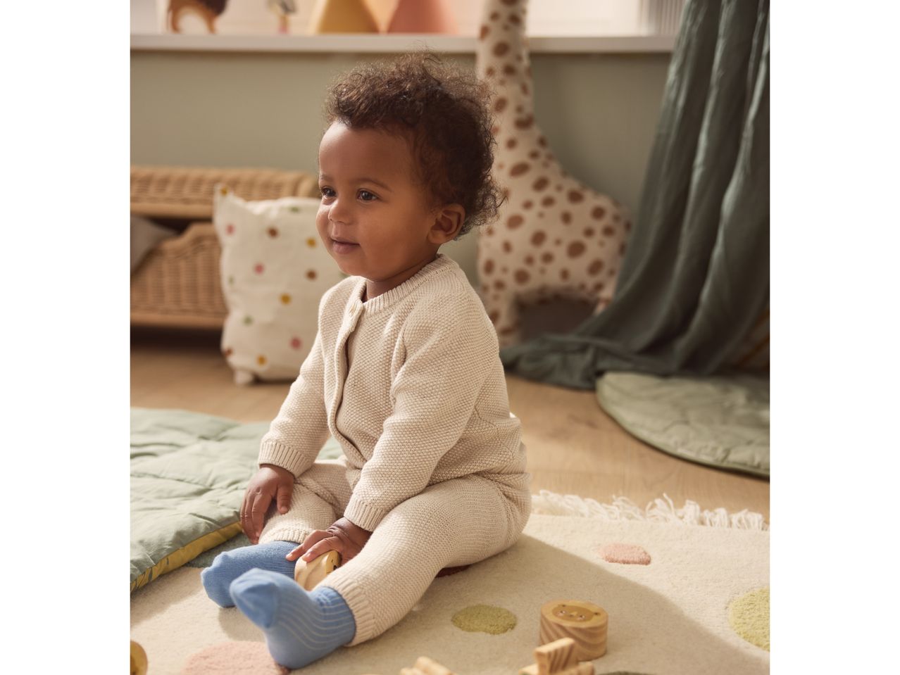 Happy baby in a knitted outfit and blue socks playing with wooden toys on a rug.